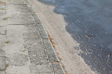 Stary, spękany chodnik z betonowych płyt wraz z krawężnikiem i fragmentem asfaltowej popękanej jezdni.