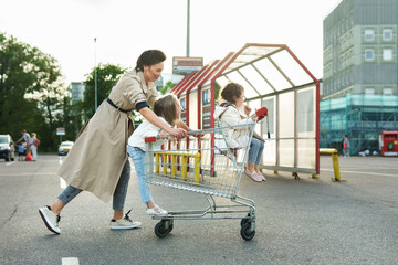 Happy mother and her daughters are having fun with a shopping cart on a parking lot beside a supermarket. © blackday