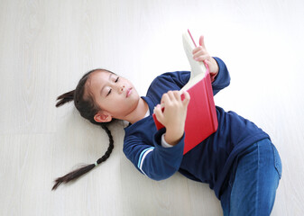 Asian little girl in casual school uniform reading a book and lying on wood laminate flooring....