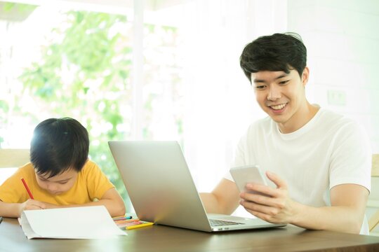 Father Working With Laptop And Mobile Phone While Son Is Drawing, Both On The Same Table. 