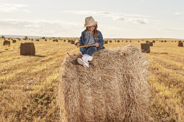 Naklejka na ściany i meble Reading girl sits over the haystack roll on field in countryside. High quality photo