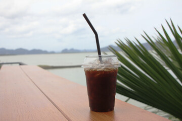 Americano black coffee is laid out on a wooden floor, behind a view of the sea and mountains in the summer under the white clouds.