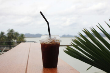 Americano black coffee packed in a clear glass placed on a red wooden floor with a natural background of the sea and mountains.