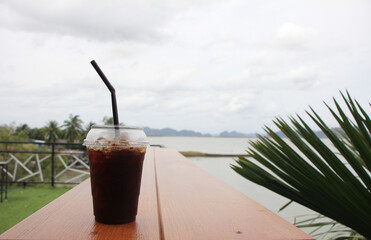 Americano black coffee is laid out on a wooden floor, behind a view of the sea and mountains in the summer under the white clouds.