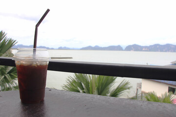 Americano black coffee is laid out on a wooden floor, behind a view of the sea and mountains in the summer under the white clouds.