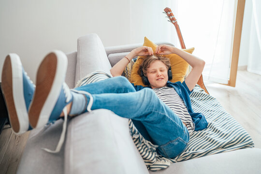 Smiling Preteen boy lying at home living room filled with sunlight on cozy sofa dressed casual jeans and sneakers listening to music using wireless headphones. Teens free time spending concept