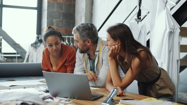 Arc Shot Of Professional Male Designer And Two Multiethnic Female Dressmakers Talking And Then Posing Together For Camera While Working In Team In Tailoring Studio