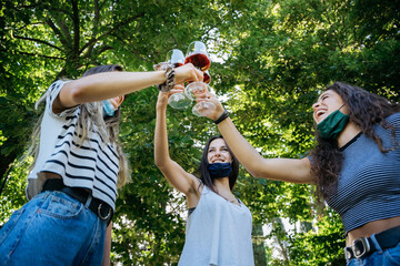 Young millennial women toasting in a park with red wine glasses in a tasting - Group of people having fun together with face mask lowered in protection by Corona virus, Covid-19