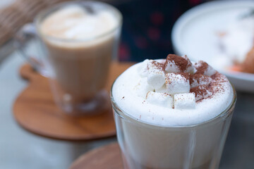 Hot chocolate with marshmallows. Hot Cappuccino in a tall class with Coffee and Croissant on background. Cappuccino or Coffee or Chocolate or Cocoa with marshmallow on top. 