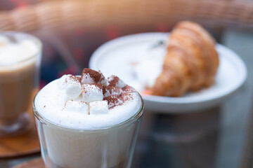 Hot chocolate with marshmallows. Hot Cappuccino in a tall class with Coffee and Croissant on background. Cappuccino or Coffee or Chocolate or Cocoa with marshmallow on top. 