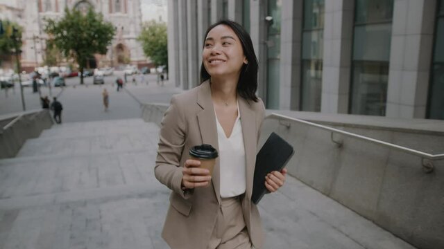 Cheerful young attractive formal lady of mixed-race ethnicity drinking coffee on office breeak walking in beautiful modern business district. People and career.