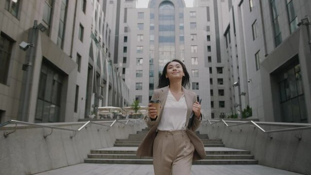 Successful young beautiful asian woman with tablet and coffee cup walking between business buildings and smiling with joy. Attractive business girl. Career people.