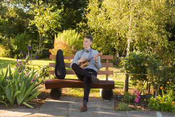 Fototapeta premium Young man sitting on bench in the park and playing ukulele.