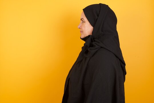 Beautiful middle aged muslim woman wearing black hijab over yellow background looking to side, relax profile pose with natural face with confident smile.