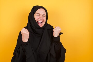 Middle aged muslim woman wearing black hijab over yellow background celebrating surprised and amazed for success with arms raised and eyes closed. Winner concept.