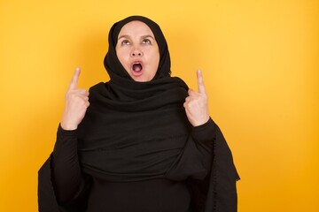 Middle aged muslim woman wearing black hijab over yellow background amazed and surprised looking up and pointing with fingers and raised arms.