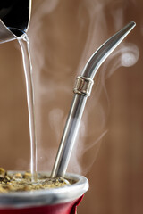 Vertical Close-Up Of Hot Traditional South American, Caffeine-Rich Infused Drink Mate Tea (Yerba Mate). Kettle Serves Hot Water Into Steaming Mate Infusion In A Modern Ceramic Cup (Mate).