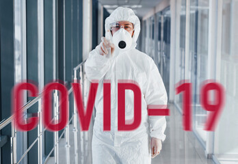 Red COVID-19 word. Nurse in protective uniform, mask and glasses standing indoors