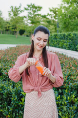 Fototapeta premium Young happy woman opens a bottle with freshly squeezed orange juice. Summer day outdoors in the park. Fresh Orange or carrot juice.