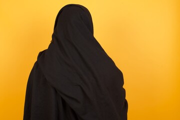 The back view of a Middle aged muslim woman wearing black hijab over yellow background Studio Shoot.