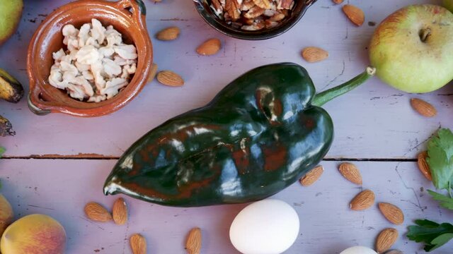 Ingredients for chile en nogada on table with poblano pepper, walnuts, bananas, pomegranates, apples, pears, parsley, almonds, eggs and peaches, panning footage.