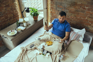 Excited cheering of favourite sport team, drinks beer. Lazy man living in his bed surrounded with messy. No need to go out to be happy. Using gadgets, watching movie and series, emotional. Fast food.