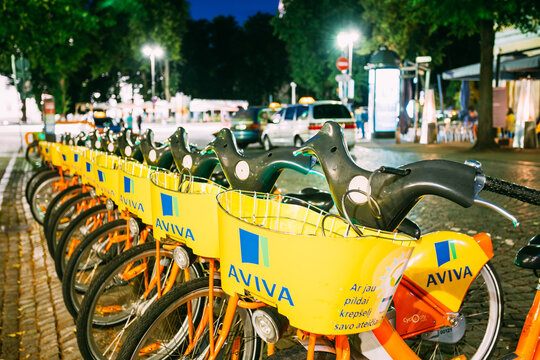 Vilnius Lithuania. Row Of Bicycles Aviva For Rent At Lit Bike Parking On Wet Pilies Street, Night
