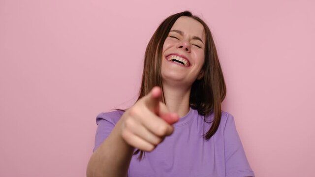 Amused happy young woman isolated on pink studio background pointing finger at camera and laughing out loud, holding belly with hilarious laughter, making fun of ridiculous appearance, wears t-shirt.