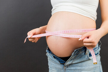 Cropped image of expecting mother in unzipped jeans measuring her pregnant belly with a tape measure at colorful background. entimeter measurement