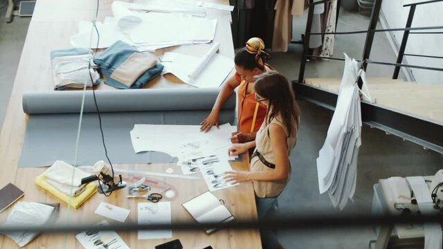 High angle shot of two multiethnic female designers discussing fashion illustrations while working together at table in tailoring studio