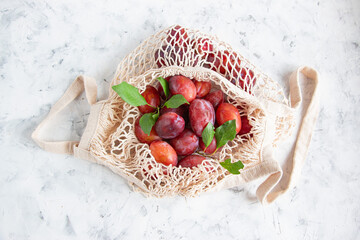 A bunch of ripe plums and a twig with leaves lie in a reusable mesh bag on a light concrete background. Fruit still life with copy space for text. Top view.