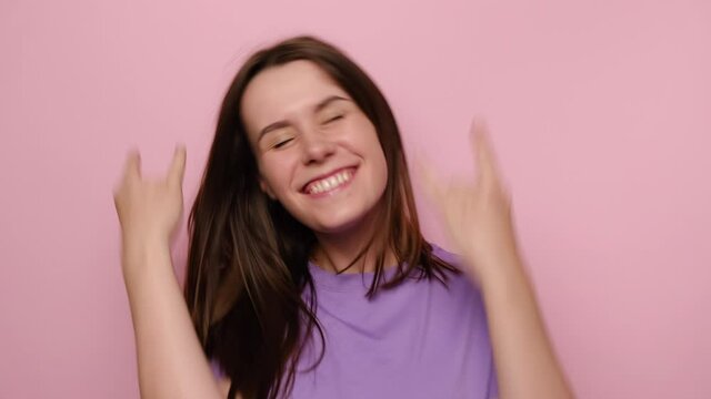 Portrait of cheerful young woman makes rock n roll sign, yells loudly, look at camera, wears purple t-shirt, devil horns hand sign gesture, stands over pink studio background, feels self confident