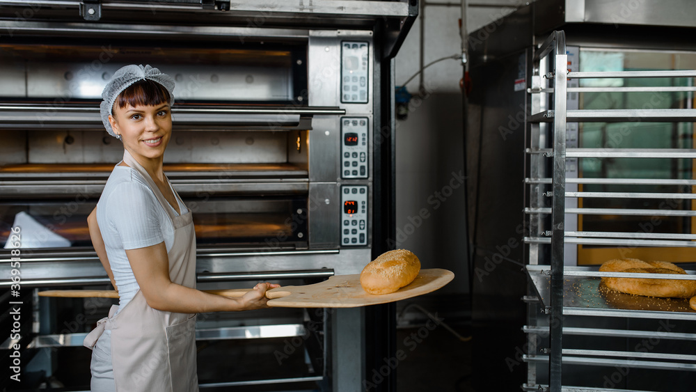 Wall mural young caucasian woman baker is holding a wood peel with fresh bread near an oven at baking manufactu