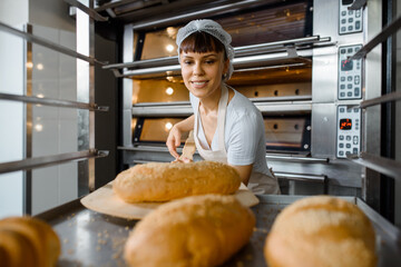 Close up of young caucasian woman baker putting the fresh bread on the shelves/rack at baking manufacture factory.