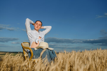 Naklejka na ściany i meble Young woman in casual outfit and a hat is sitting on a vintage chair in the wheat field.