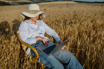 Naklejka na ściany i meble Young woman freelancer is working on a computer outdoors in the wheat field. She is sitting on a vintage chair and enjoying the ability to work from anywhere