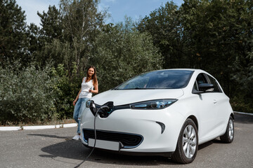 Young beautiful woman is charging her electric car at the charging station situated in the forest.
