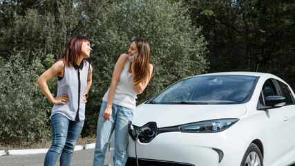 Two happy young beautiful women are talking to each other until their electric car is charging at the charging station situated in the forest.