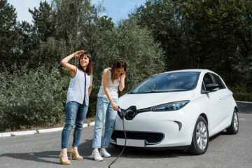 Two happy young beautiful women are talking to each other until their electric car is charging at the charging station situated in the forest.