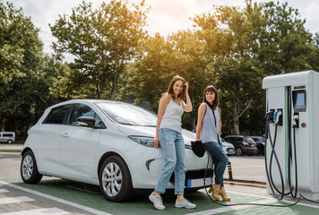 Two happy young beautiful women are talking to each other until their electric car is charging at...