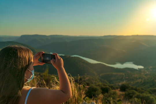 Blonde girl with surgical mask by the covid-19 takes a nice picture with her smartphone from the top of the valley while it is getting dark with the Douro river and the mountains