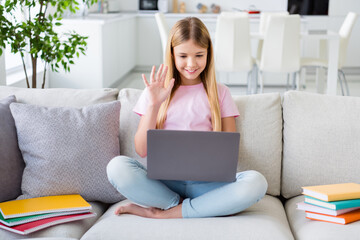 Full length photo of little kid girl sit cozy couch legs crossed have online lesson conversation greet teacher wave hand use laptop study remote in house indoors