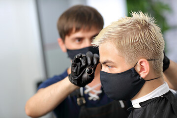 A Barber makes cuts the hair of a young guy in a protective mask. A Barber in black nitrile gloves...