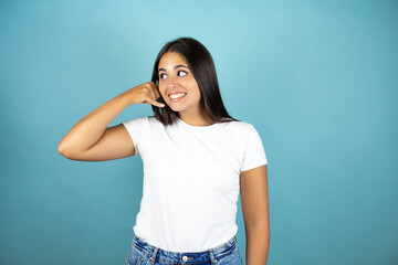 Young beautiful woman over isolated blue background looking side and smiling doing phone gesture with hand and fingers like talking on the telephone