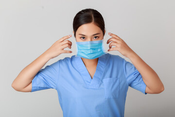 Young attractive Asian practitioner in blue hospital uniform wearing protection face mask with smiley face isolated on white, health care during pandemic crisis.