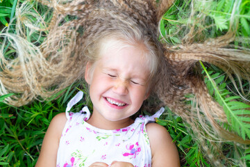 A little Caucasian girl with long blond hair in a white sundress lies on her back in the grass, closed her eyes, smiles, laughs. Childhood, happiness, reunion with nature, lifestyle. Close-up portrait
