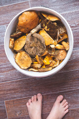 Plastic white bucket with forest mushrooms, close-up top view. Child's bare feet. Autumn harvest, gifts of the forest.