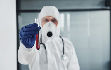 Male doctor scientist in lab coat, defensive eyewear and mask holds test tube with blood