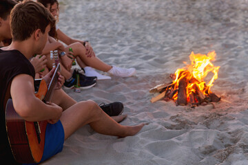 Group of friends sitting with guitar on the beach near the campfire. Teenagers have fun on the vacation. Boy playing guitar and singing song with his friends near the campfire.