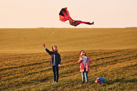 Two little girls friends have fun together with kite and toy car on the field at sunny daytime